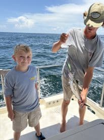 A young boy smiles proudly alongside a teenager holding a freshly caught fish while on a fishing charter in open waters.
