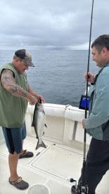 Two anglers aboard a boat, one holding a freshly caught fish while the other prepares to assist. The scene captures the excitement of fishing on open water.