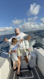 A young boy and an adult are engaged in fishing on a boat, with the adult assisting the boy as he reels in a catch, showcasing a lively fishing experience.