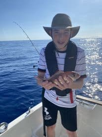 A young angler proudly displaying a Snapper while fishing on a sunny day in open waters.