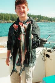 A young angler proudly displaying multiple freshly caught salmon aboard a fishing boat in a scenic coastal setting.