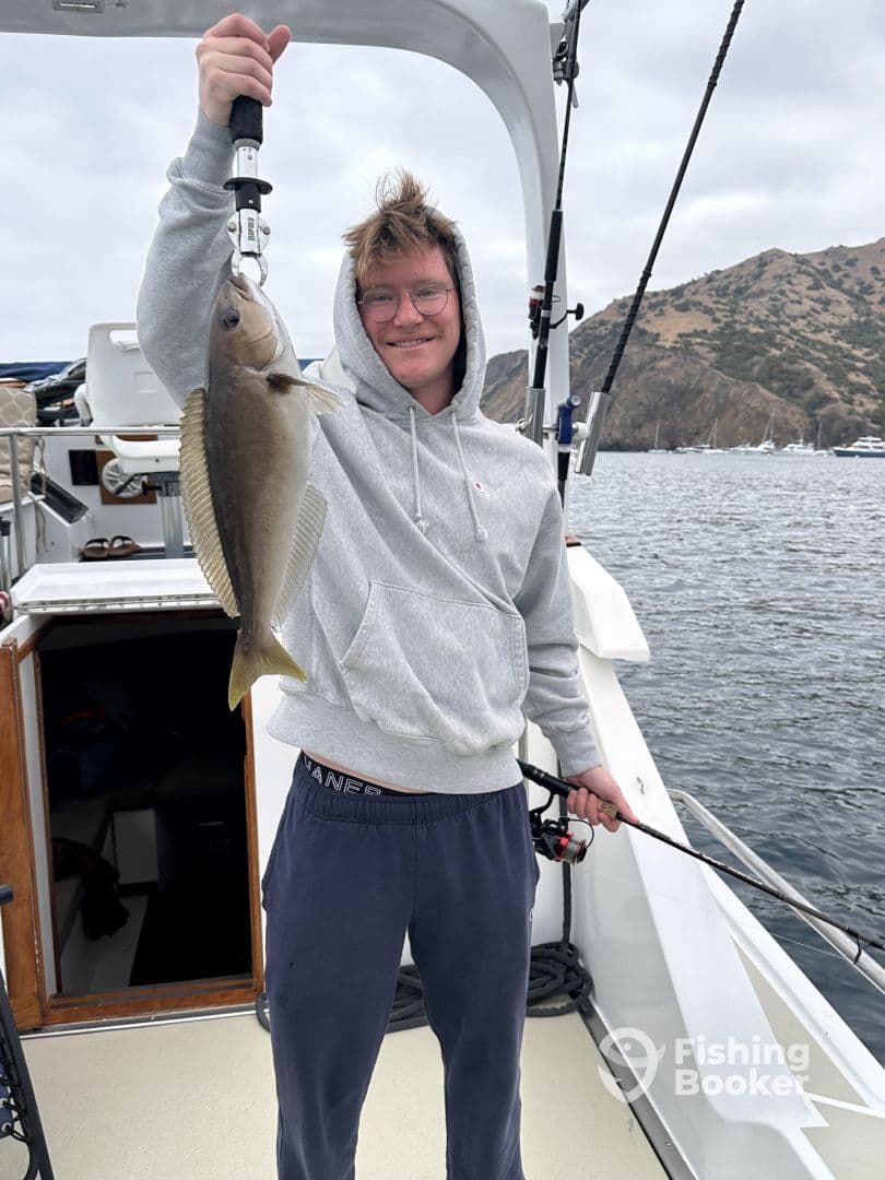 A young angler proudly displaying a Halibut caught while fishing off the coast, with a scenic backdrop of rocky hills.