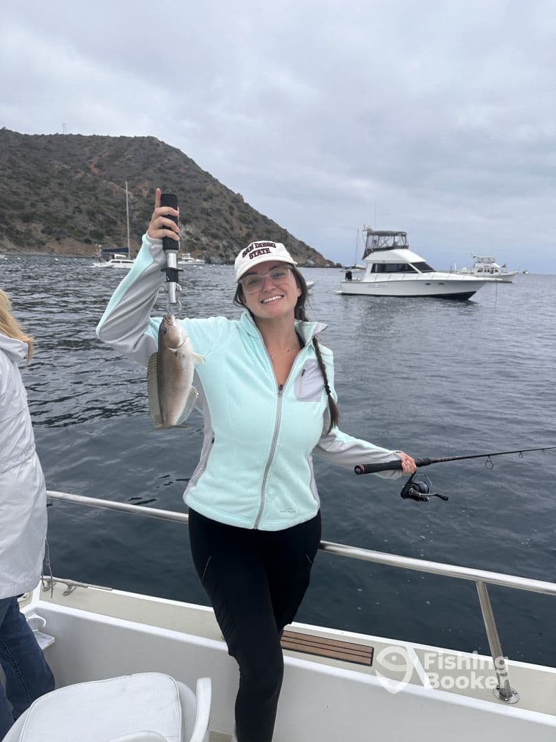 A happy angler displaying her catch while fishing off the coast, with boats and a scenic backdrop in the background.