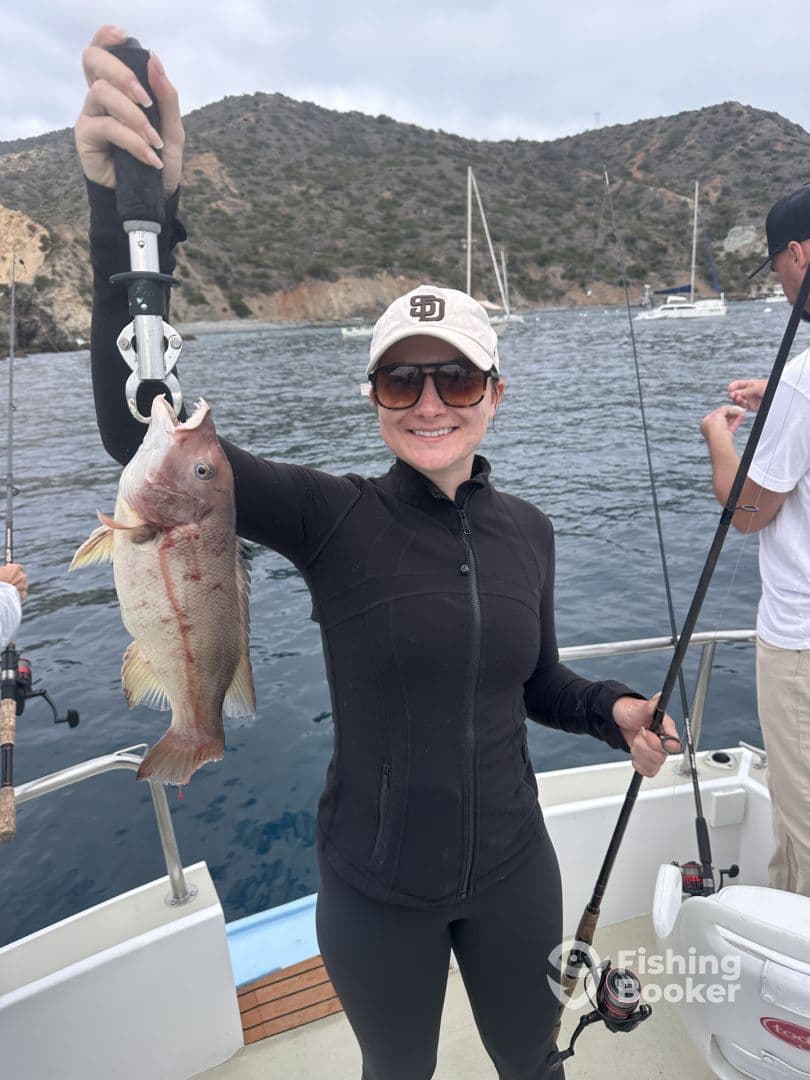 An angler proudly displaying a freshly caught fish while fishing off the coast, surrounded by a scenic backdrop of hills and boats.