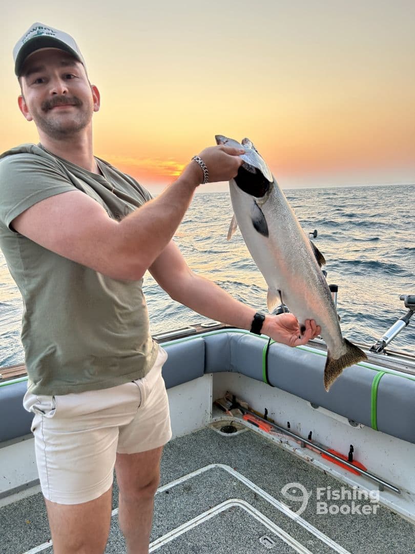 An angler proudly displaying a large Salmon against a stunning sunset backdrop while on a fishing trip.