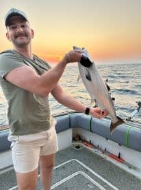 An angler proudly displaying a large Salmon against a stunning sunset backdrop while on a fishing trip.