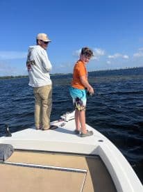 A young angler is actively fishing from the bow of a boat, enjoying a sunny day on the water with an adult companion.