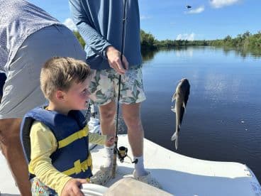 A young boy in a life jacket watches excitedly as a catfish is reeled in during a family fishing trip on calm waters.