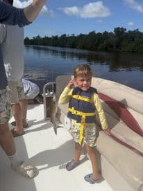 A young boy wearing a life jacket proudly displays his catch while fishing on a boat in a calm river setting.