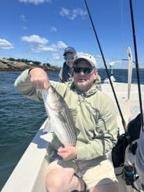 An adult angler proudly displaying a striped bass while fishing on a boat, with a young companion in the background enjoying the day on the water.