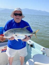 An older angler proudly displaying a large Jack Crevalle while fishing on a sunny day in a coastal setting.
