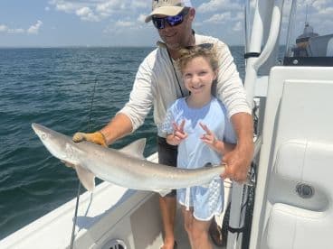 A young girl and an adult proudly display a caught shark while on a fishing trip, showcasing a fun moment on the water.