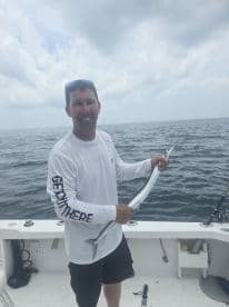 An angler proudly displaying a long, slender fish, likely a barracuda, aboard a fishing boat in open waters.