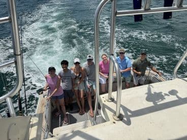 A group of six anglers enjoying their time on a fishing boat, with rods in hand and the ocean in the background, showcasing a lively fishing atmosphere.