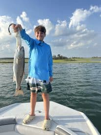 A young angler proudly displaying a large Speckled Trout while standing on the bow of a boat in a scenic fishing environment.