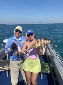 Two anglers proudly displaying a large Grouper caught while fishing on a boat in clear blue waters.