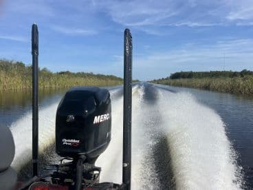 A view from the back of a boat, showcasing the powerful Mercury engine as it speeds through a calm waterway surrounded by lush vegetation.