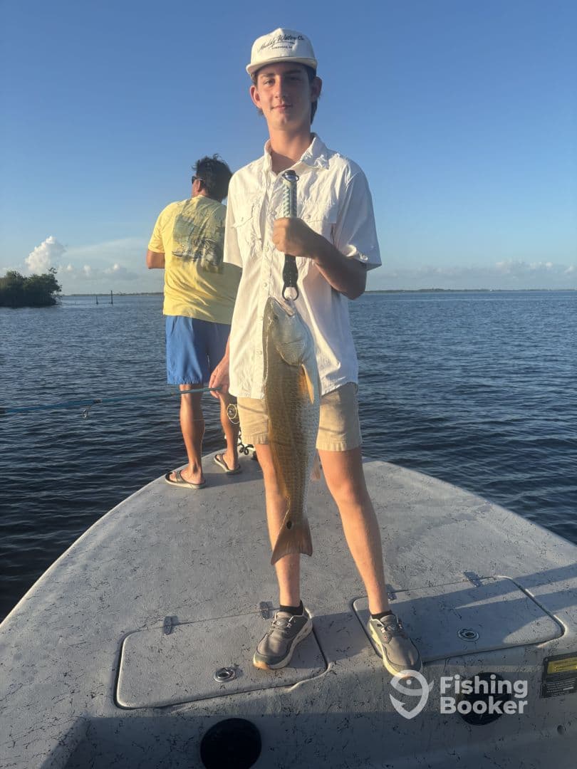 A young angler proudly displaying a Redfish while fishing on a boat in a scenic coastal environment.