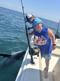 A young angler gives a thumbs up while fishing, with a shark visible in the water beside the boat, showcasing an exciting moment on a deep-sea fishing trip.