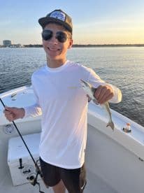 A young angler proudly displaying a small fish while fishing on a boat during sunset.
