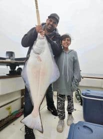 A proud angler and a young girl display a large Halibut on a fishing boat, showcasing a successful day on the water.