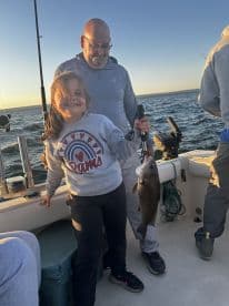 A young girl proudly displaying her catch, a sizable fish, while fishing with family on a boat during sunset.