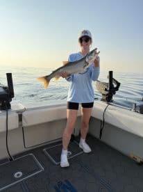 A young angler proudly displaying a large Lake Trout while fishing on a calm lake, showcasing a beautiful sunrise backdrop.