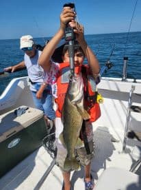A young angler proudly displaying a large Walleye while fishing on a boat, with a family member in the background.