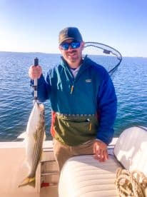 An angler proudly displaying a striped bass while fishing on a boat, showcasing a clear blue sky and calm waters.