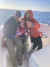A family proudly displaying a large Striped Bass on a fishing trip, with a scenic water backdrop.