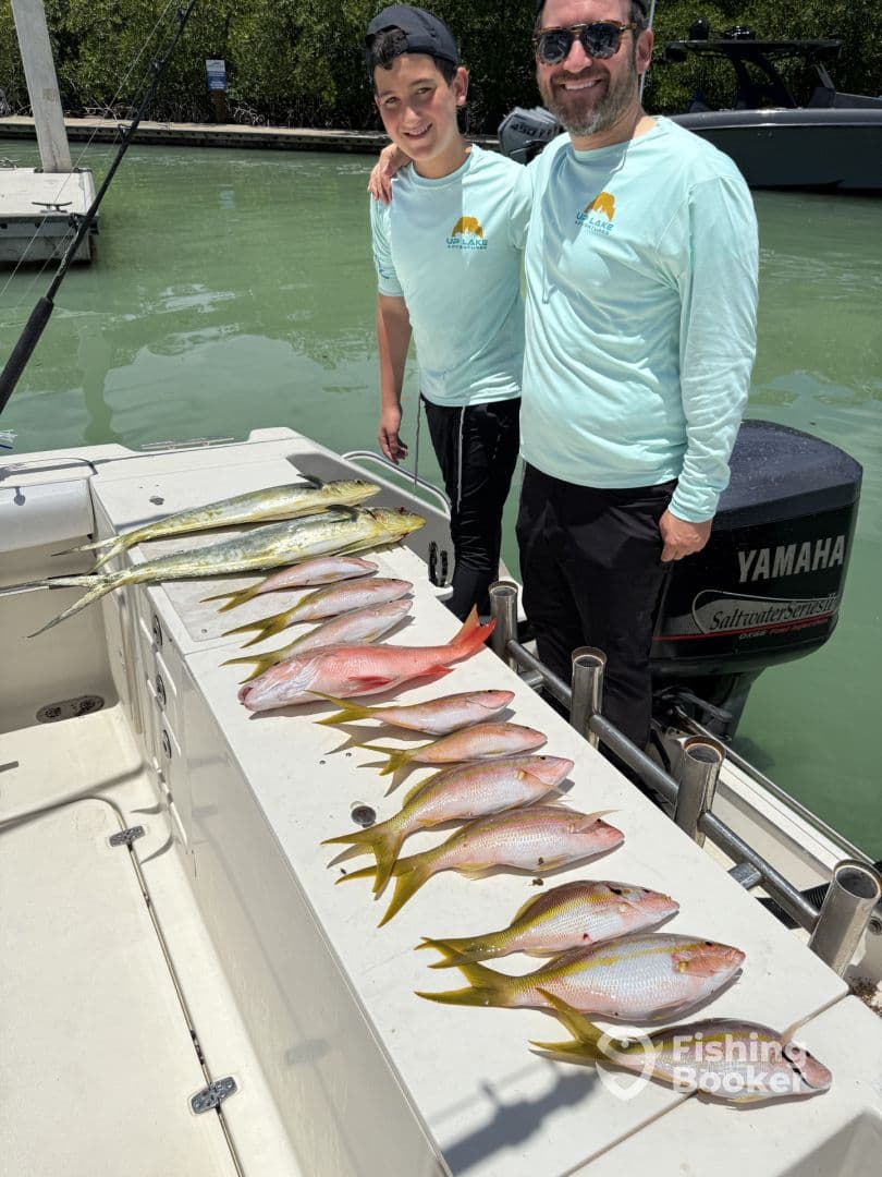 A father and son proudly display their catch of various fish, including Mahi Mahi and Snapper, on a boat after a successful fishing trip.