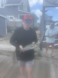 A young angler proudly displaying a caught fish at the dock, with a coastal house in the background.