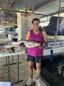 An angler proudly displaying a large striped bass at a marina, surrounded by freshly caught fish on a table.