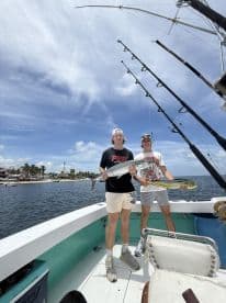 Two anglers proudly displaying their catches, a Mahi Mahi and a Barracuda, aboard a fishing boat in a tropical setting.