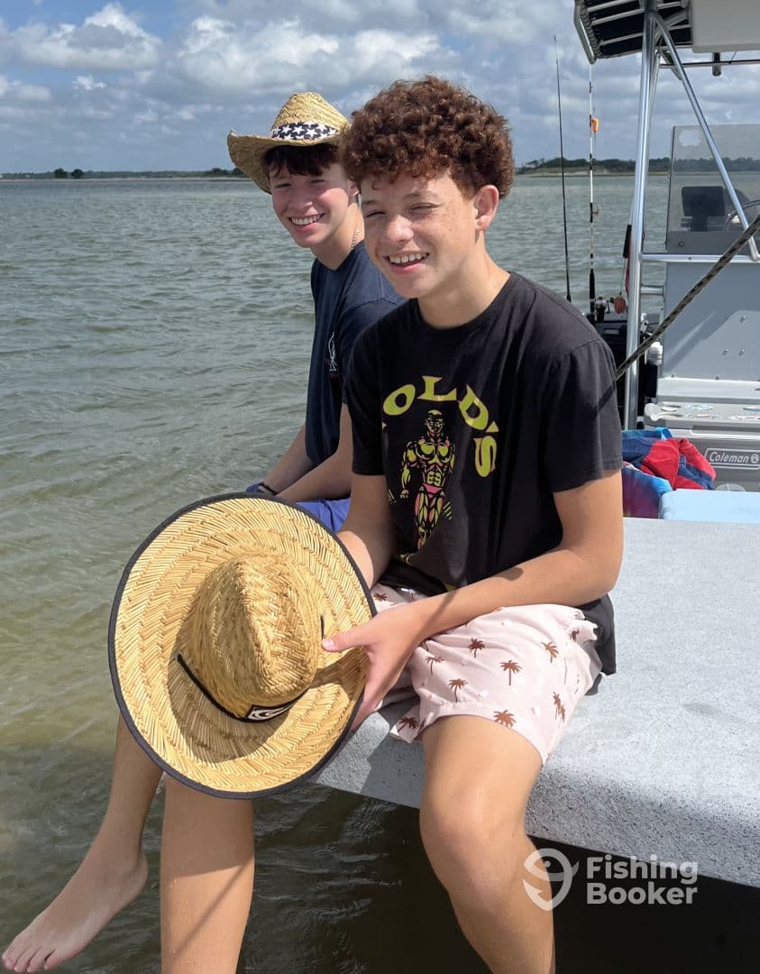 Two young anglers enjoying a day on the water, seated on the edge of a boat with fishing gear visible in the background.