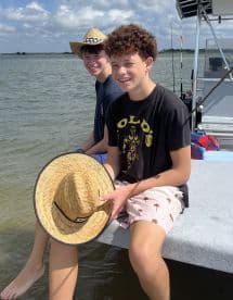 Two young anglers enjoying a day on the water, seated on the edge of a boat with fishing gear visible in the background.