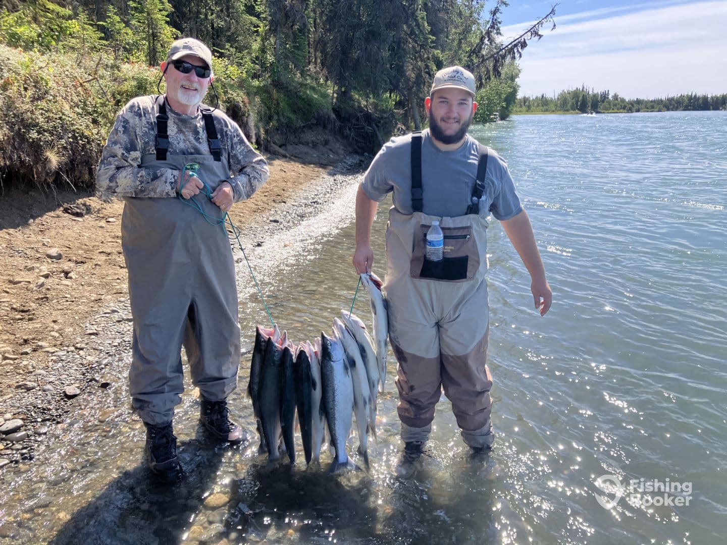Two anglers proudly displaying their catch of salmon along a riverbank, showcasing a successful fishing trip in a scenic environment.