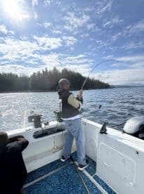 An angler actively reeling in a fish while fishing on a boat surrounded by scenic forested coastline.