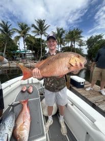 A young angler proudly displaying a large Red Snapper aboard a fishing boat, surrounded by palm trees and a scenic waterfront.