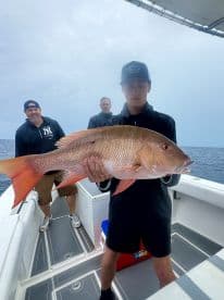 A young angler proudly displaying a large Snapper while fishing offshore, with two adults in the background.