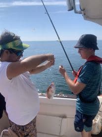 Two young anglers engaged in fishing on a boat, with one holding a freshly caught Snapper while the other assists in reeling in.