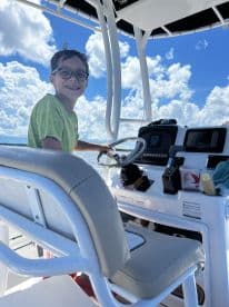 A young boy enjoying his time at the helm of a modern fishing boat, surrounded by a bright blue sky and fluffy clouds.