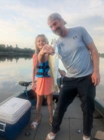 A young girl and an adult proudly displaying a Walleye caught during a peaceful morning fishing trip on a lake.