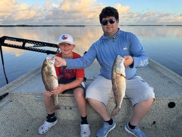 A young angler and an adult proudly displaying their catches of Redfish while enjoying a serene day on the water.