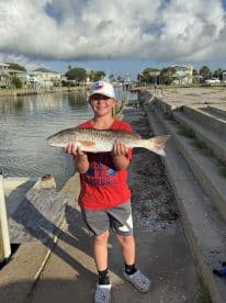 A young angler proudly displaying a Redfish caught at a coastal fishing spot, with residential homes lining the canal in the background.