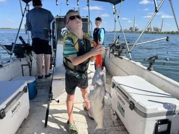 A young angler proudly holding a large catfish while fishing on a boat, with another child in the background and a scenic waterfront view.