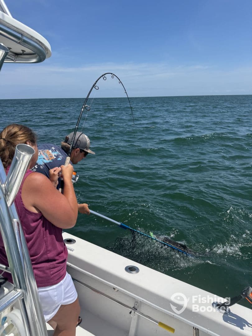 A young angler is reeling in a fish while another person prepares to net it, showcasing an exciting moment during a fishing trip on a boat.