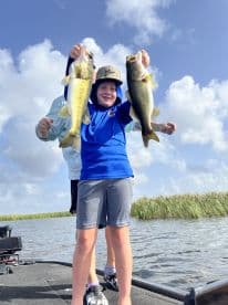 A young angler proudly displaying two large Largemouth Bass while fishing on a boat in a scenic waterway.