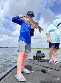A young angler proudly displaying a large Largemouth Bass while fishing on a boat, with another person in the background.
