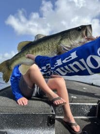 A young angler proudly displaying a large Largemouth Bass while sitting on a fishing boat, showcasing a sunny day on the water.